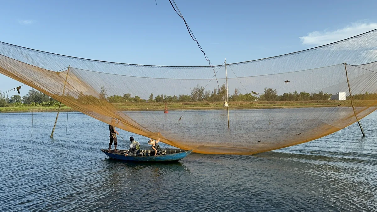 Fishermen operate a large square lift net from a small boat on a river near Hoi An's countryside.