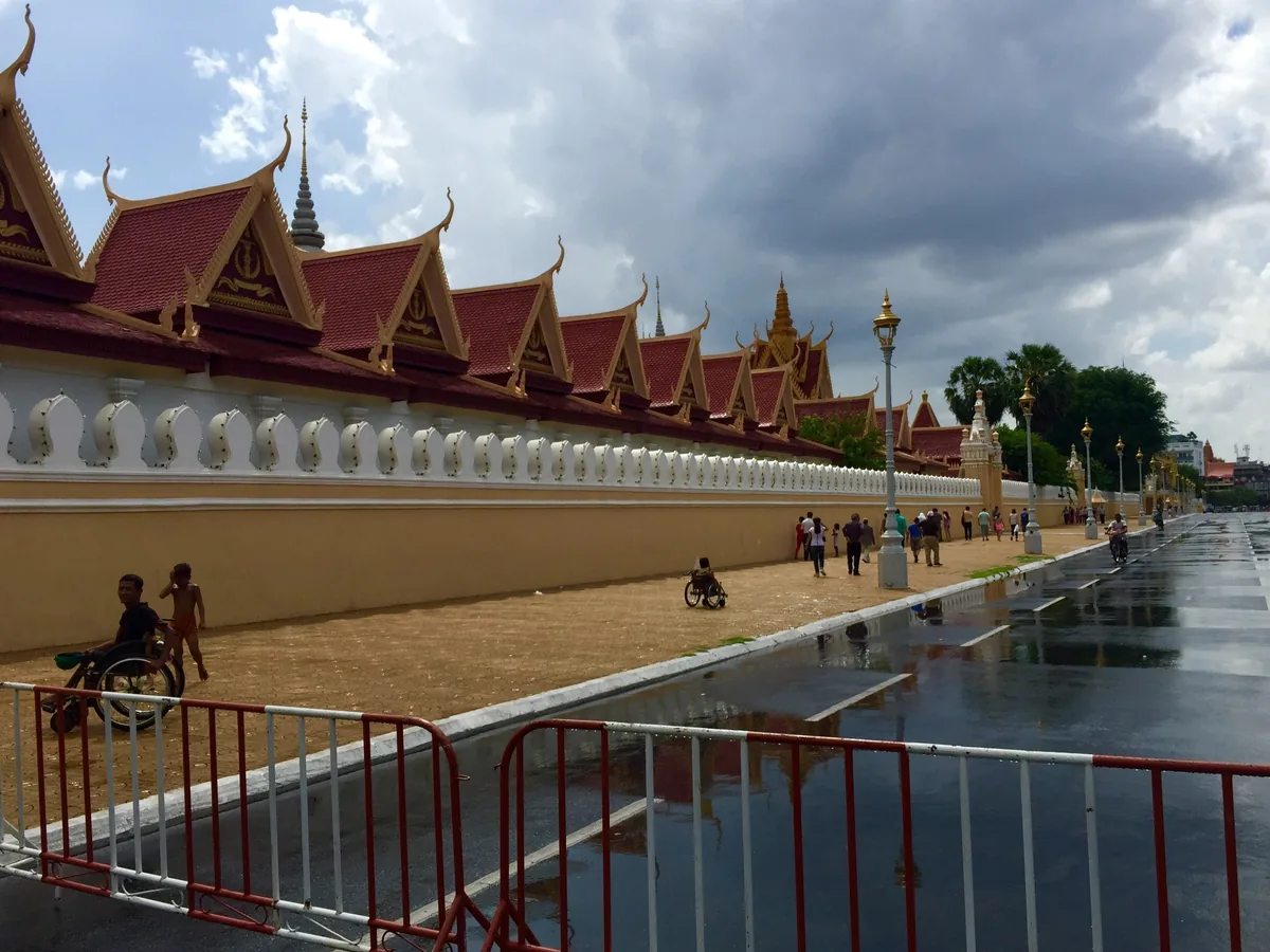 Tiered roofs of the Royal Palace wall line a wet riverside street in Phnom Penh under a cloudy sky.