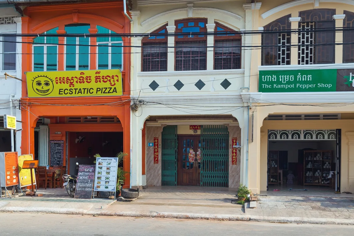 Colorful colonial shophouses line an old market street in Kampot, including the Kampot Pepper Shop and a pizza restaurant.
