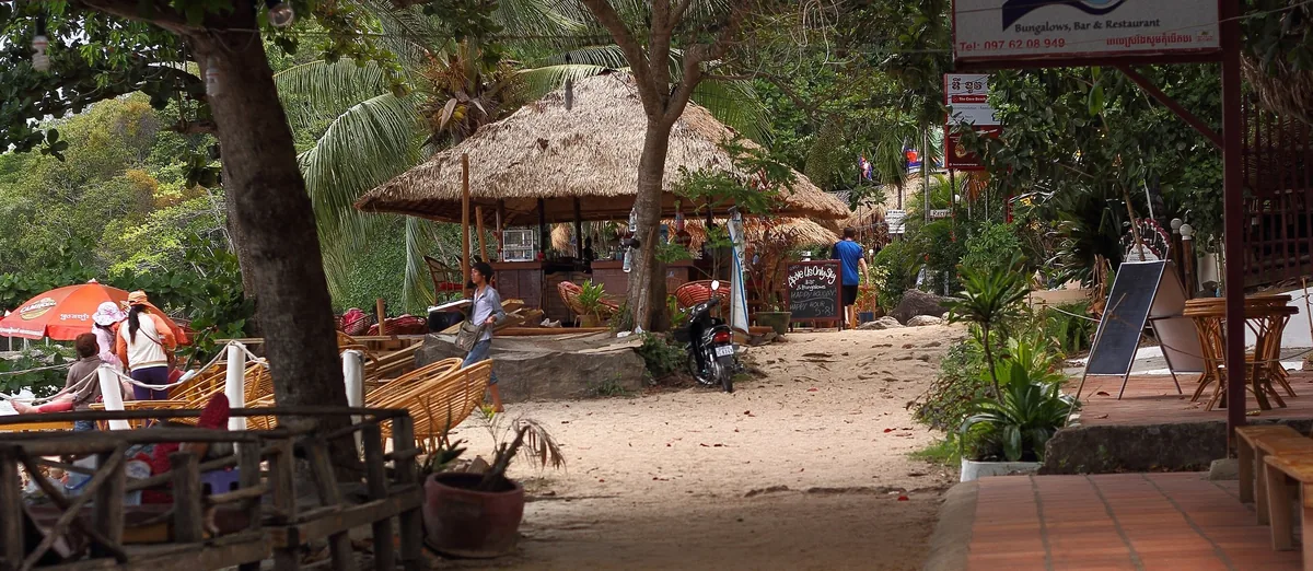 A thatched-roof beachside bar and café sits under shady trees along a sandy path in Sihanoukville, Cambodia.