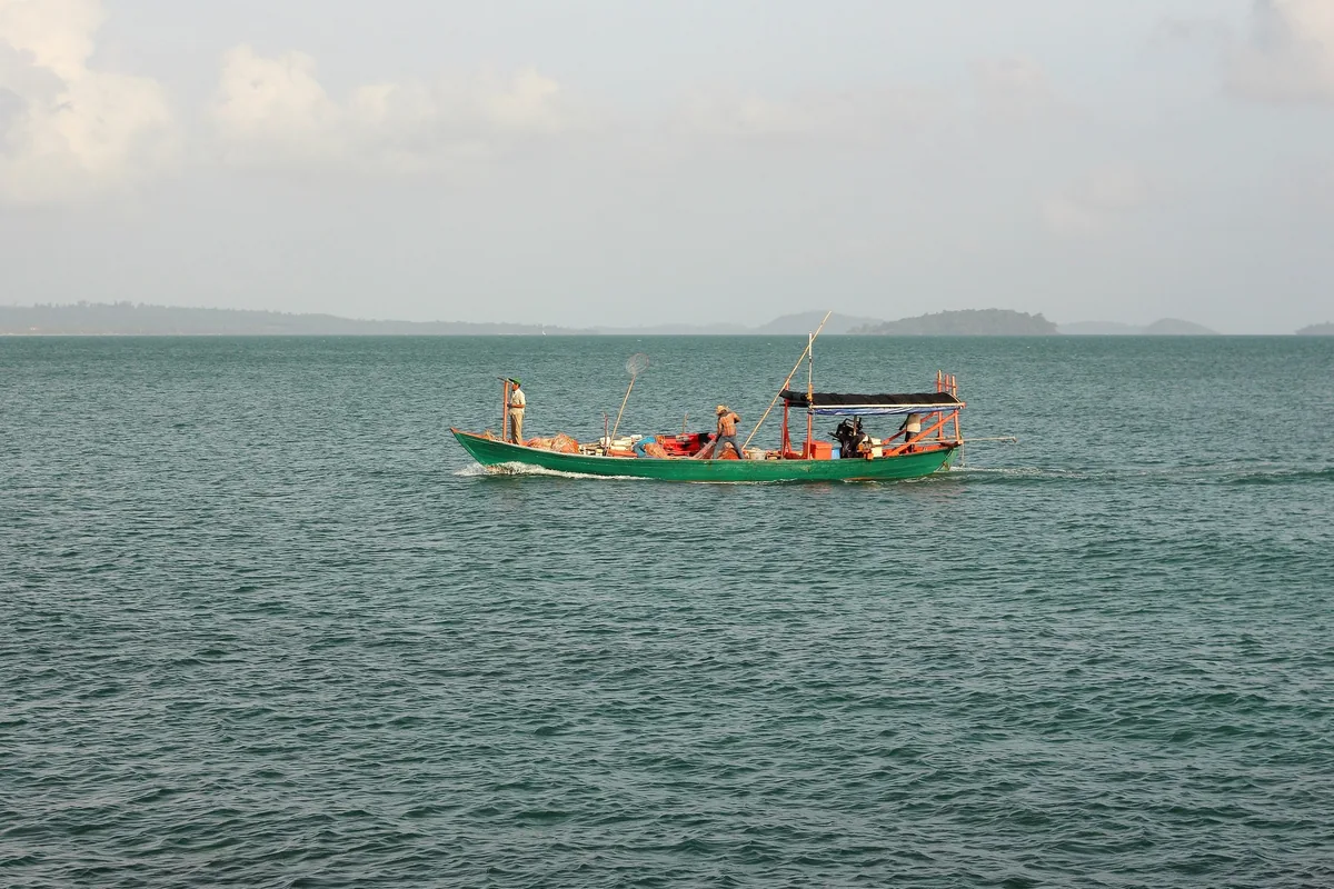 A green wooden fishing boat with two men aboard crosses calm waters near Sihanoukville, with distant islands on the horizon.