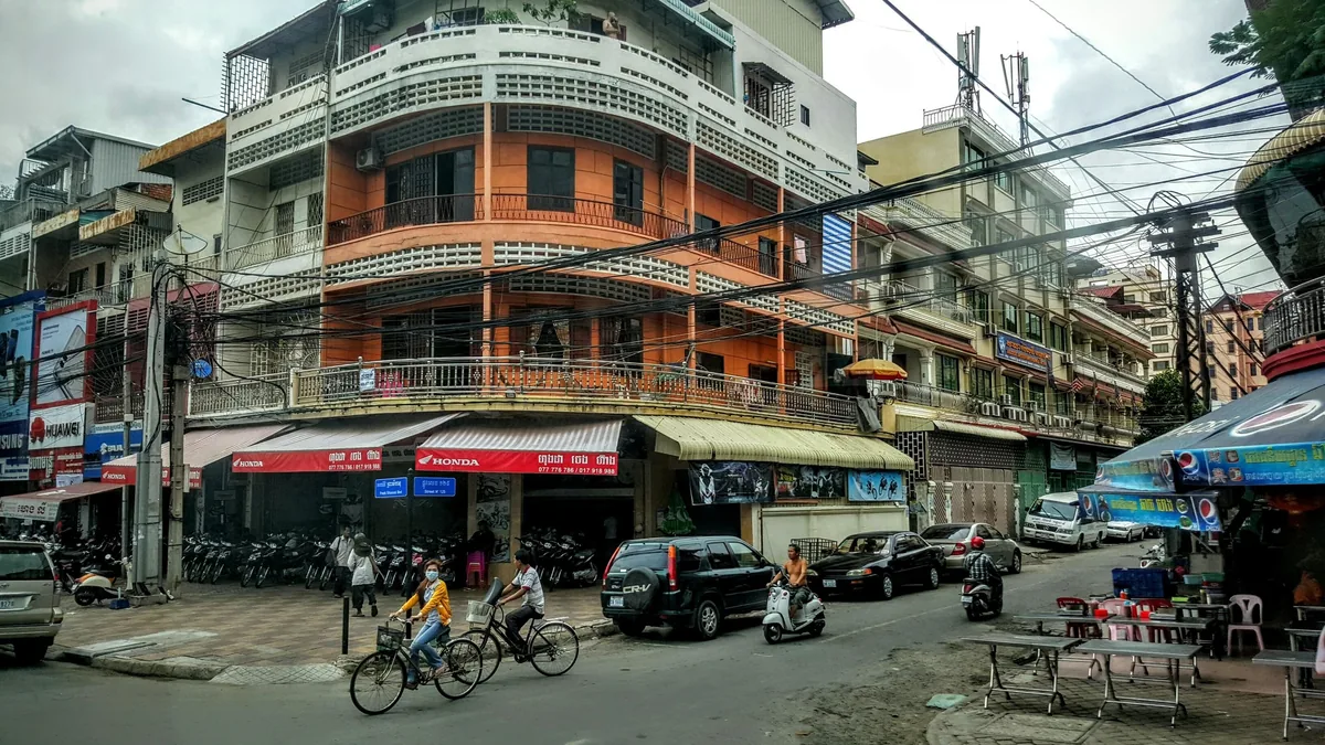 A busy street corner in Phnom Penh with cyclists, parked scooters, and an orange corner building strung with overhead wires.