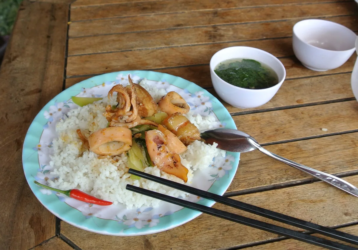 A plate of stir-fried squid with rice, chili, and a bowl of soup on a wooden table in Vietnam.