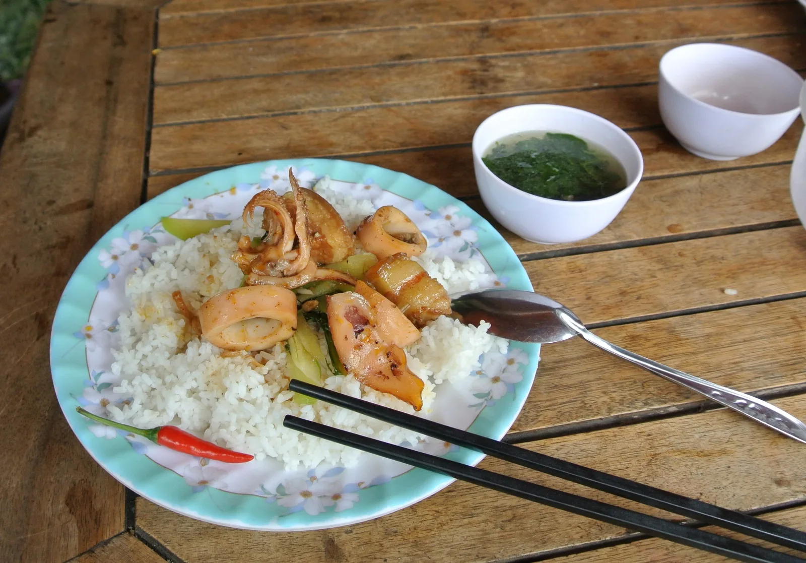 A plate of stir-fried squid with rice, chili, and a bowl of soup on a wooden table in Vietnam.