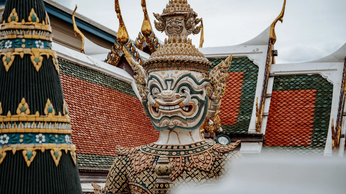 A guardian yaksha statue stands before ornate tiled temple roofs in Thailand.