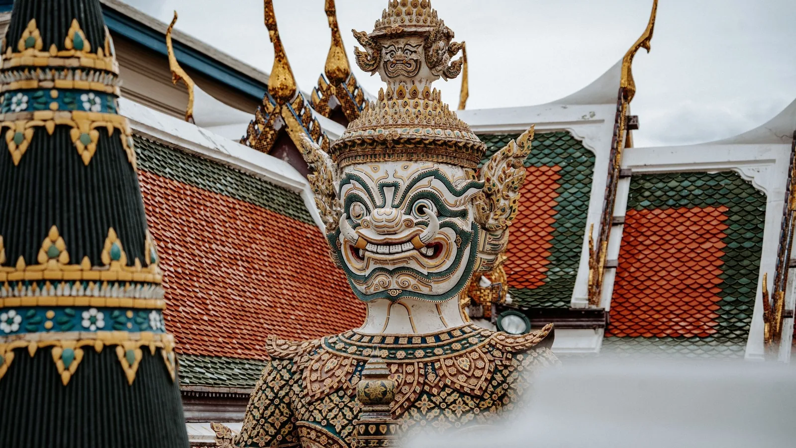 A guardian yaksha statue stands before ornate tiled temple roofs in Thailand.