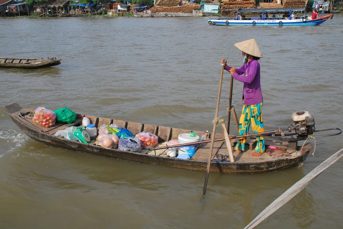 A vendor in a conical hat steers a wooden boat loaded with goods along a muddy Mekong river in Cambodia.