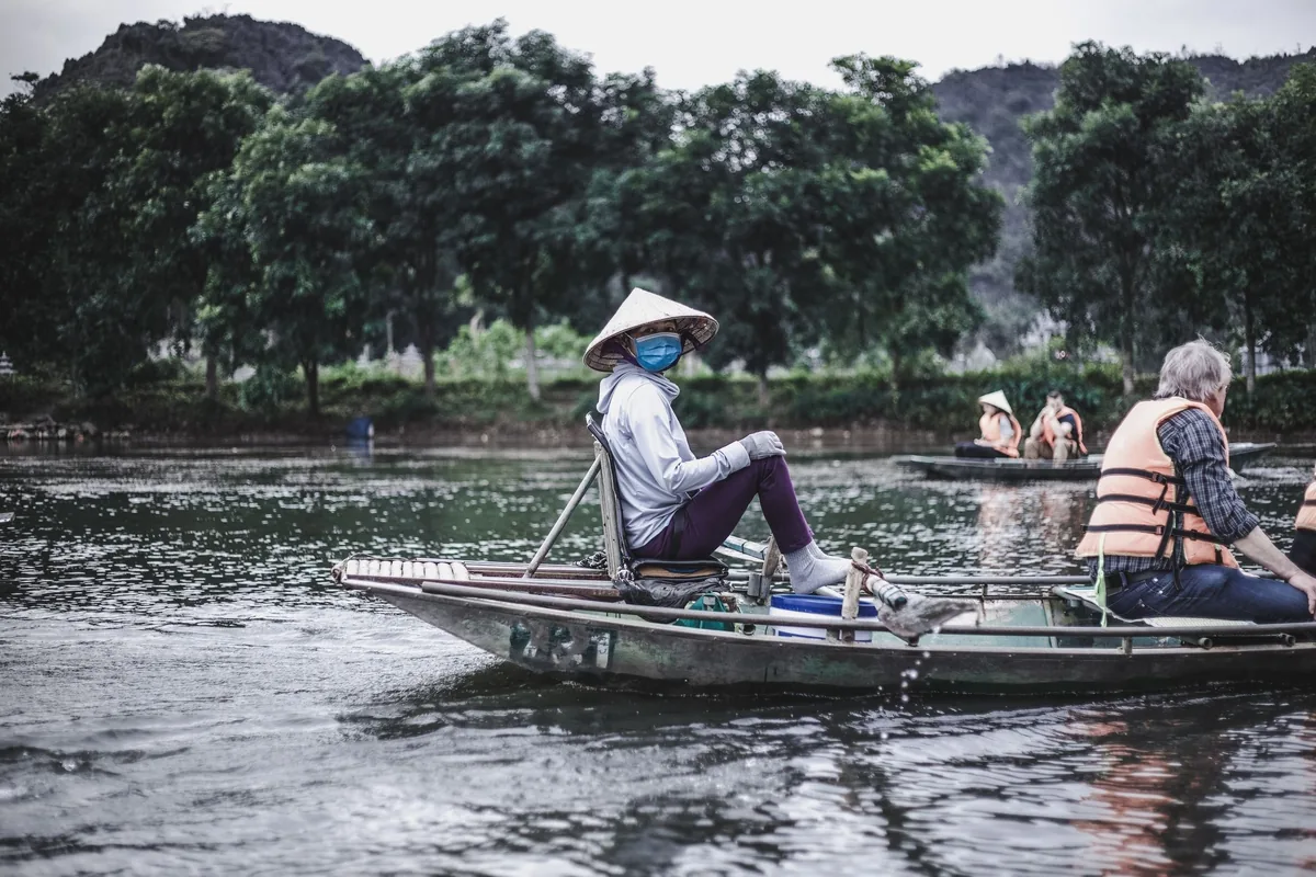 A boatman in a conical hat rows a small wooden boat with tourists on a calm river in Vietnam.