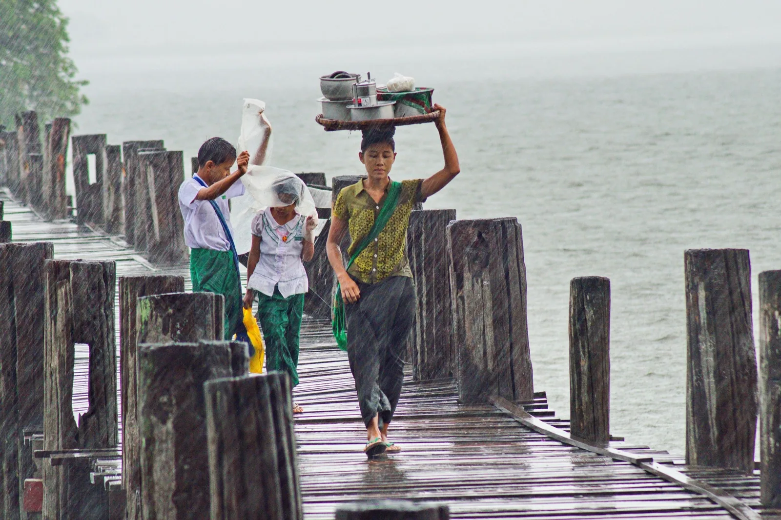 A vendor balances a tray of goods on her head while walking across U Bein Bridge in the rain, Mandalay.