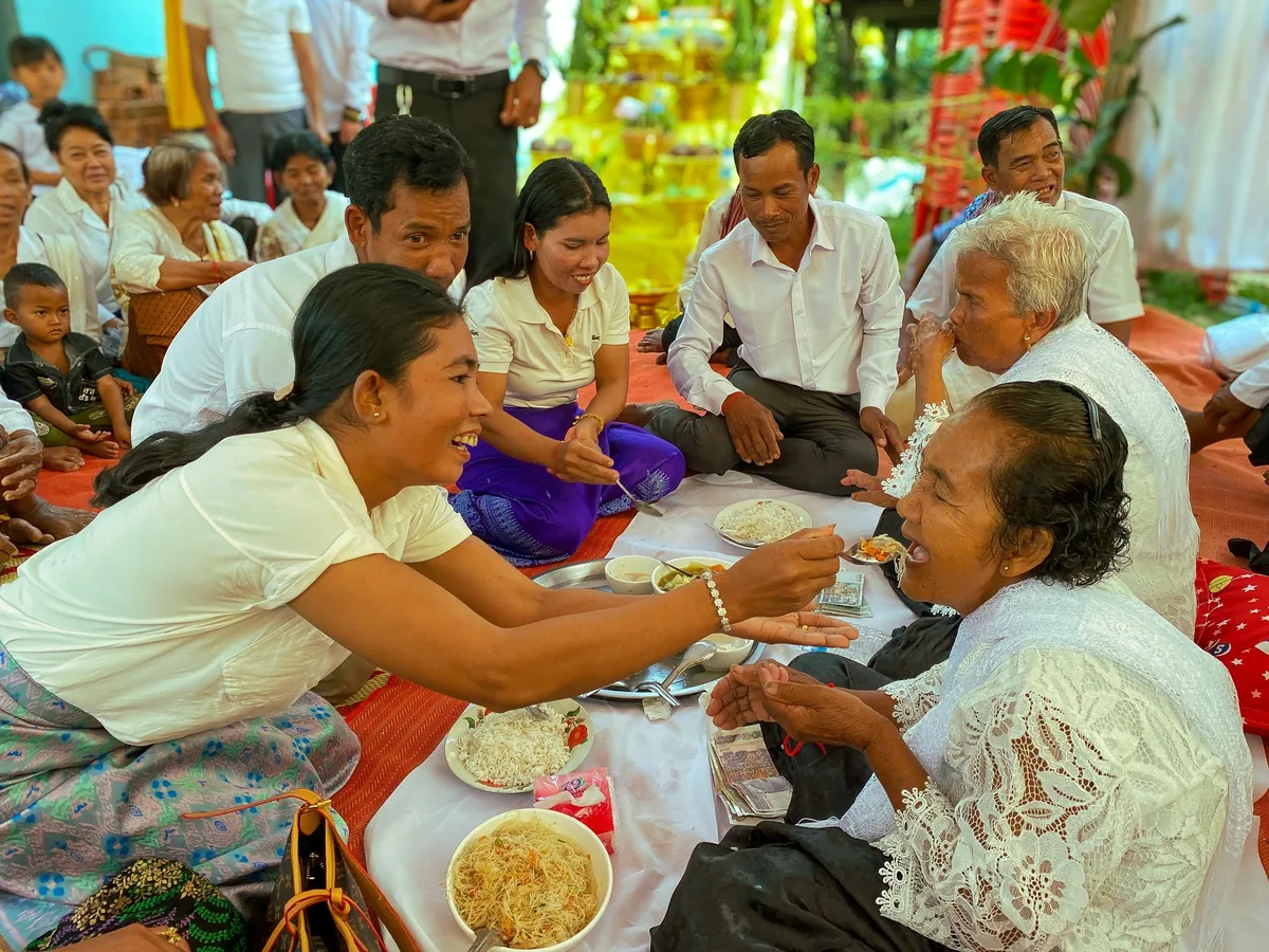 A Cambodian family sits on mats sharing a festive meal, laughing as a woman feeds an elder during a traditional gathering.