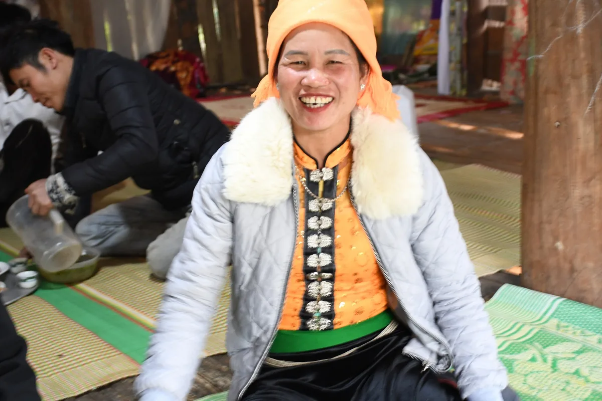 A smiling woman in traditional dress with a fur-trimmed jacket and orange headscarf sits on a mat inside a wooden house in.