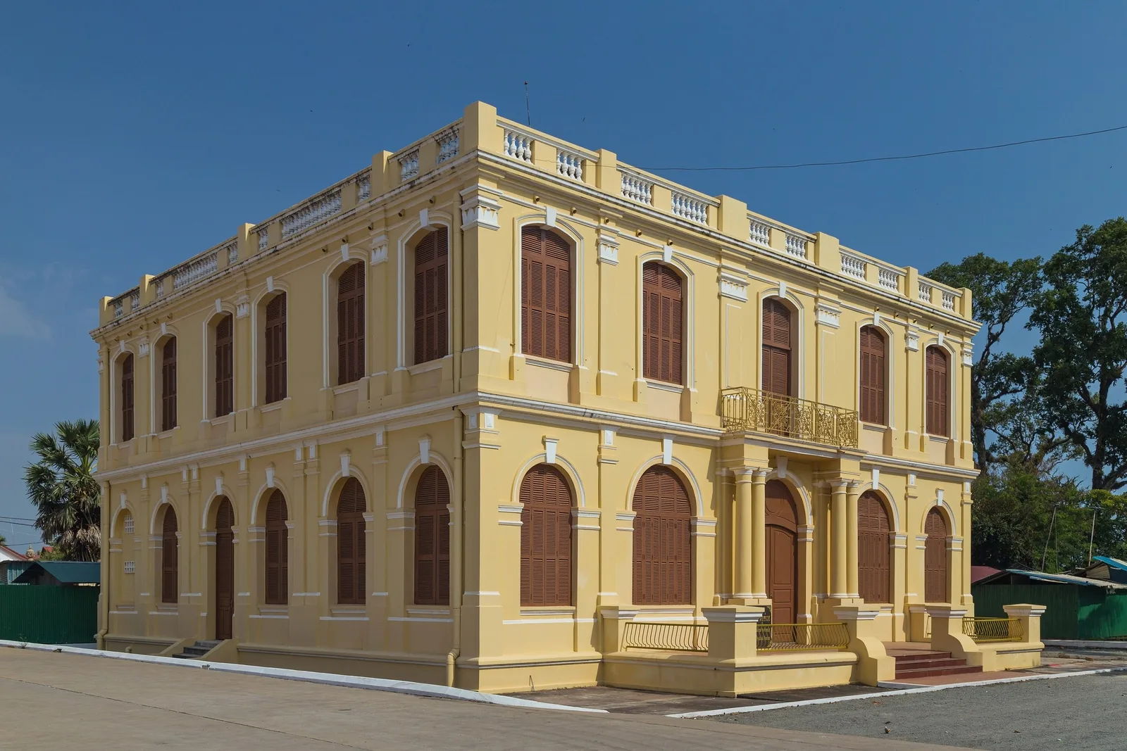 A colonial-era yellow building with arched shuttered windows stands under a clear blue sky in Kampot.