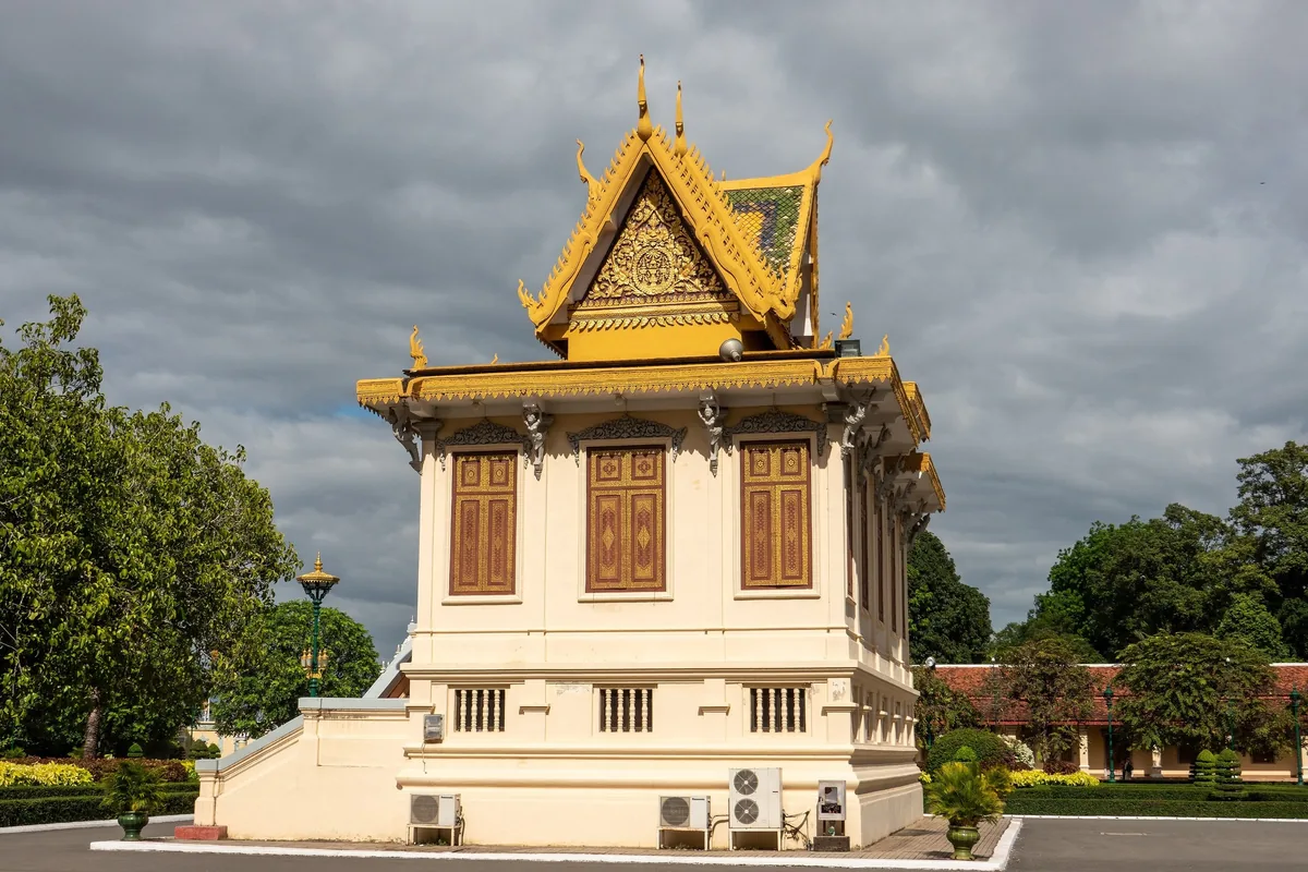Golden-roofed pavilion with ornate windows stands within the Royal Palace grounds in Phnom Penh under cloudy skies.