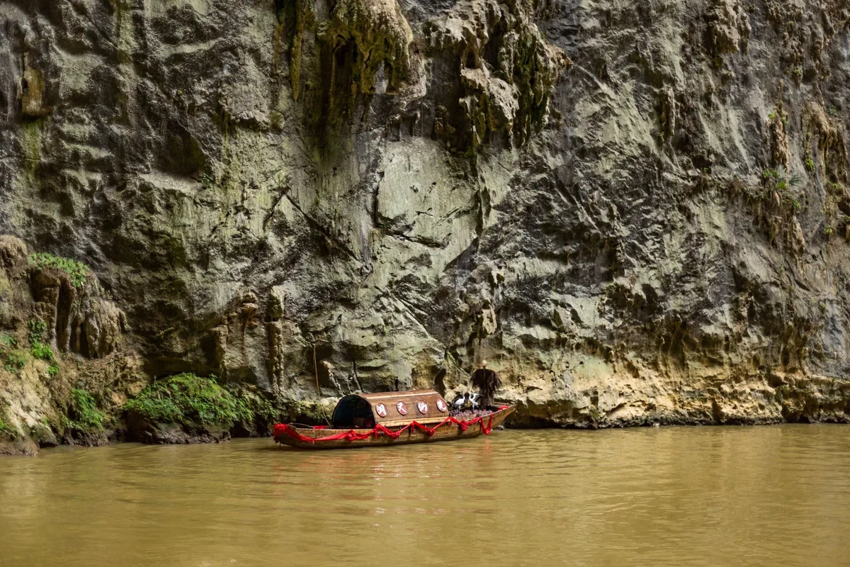 A small wooden boat glides along a river beneath a towering limestone cliff in Laos.