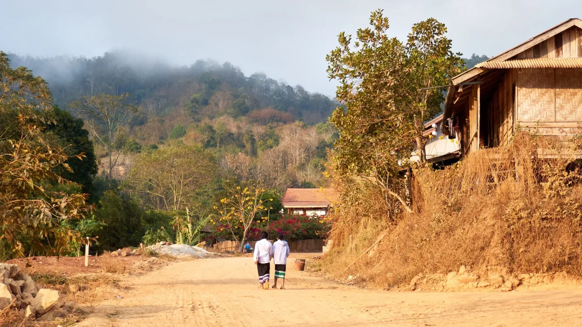 Two children walk down a dirt road through a misty village in rural Laos.