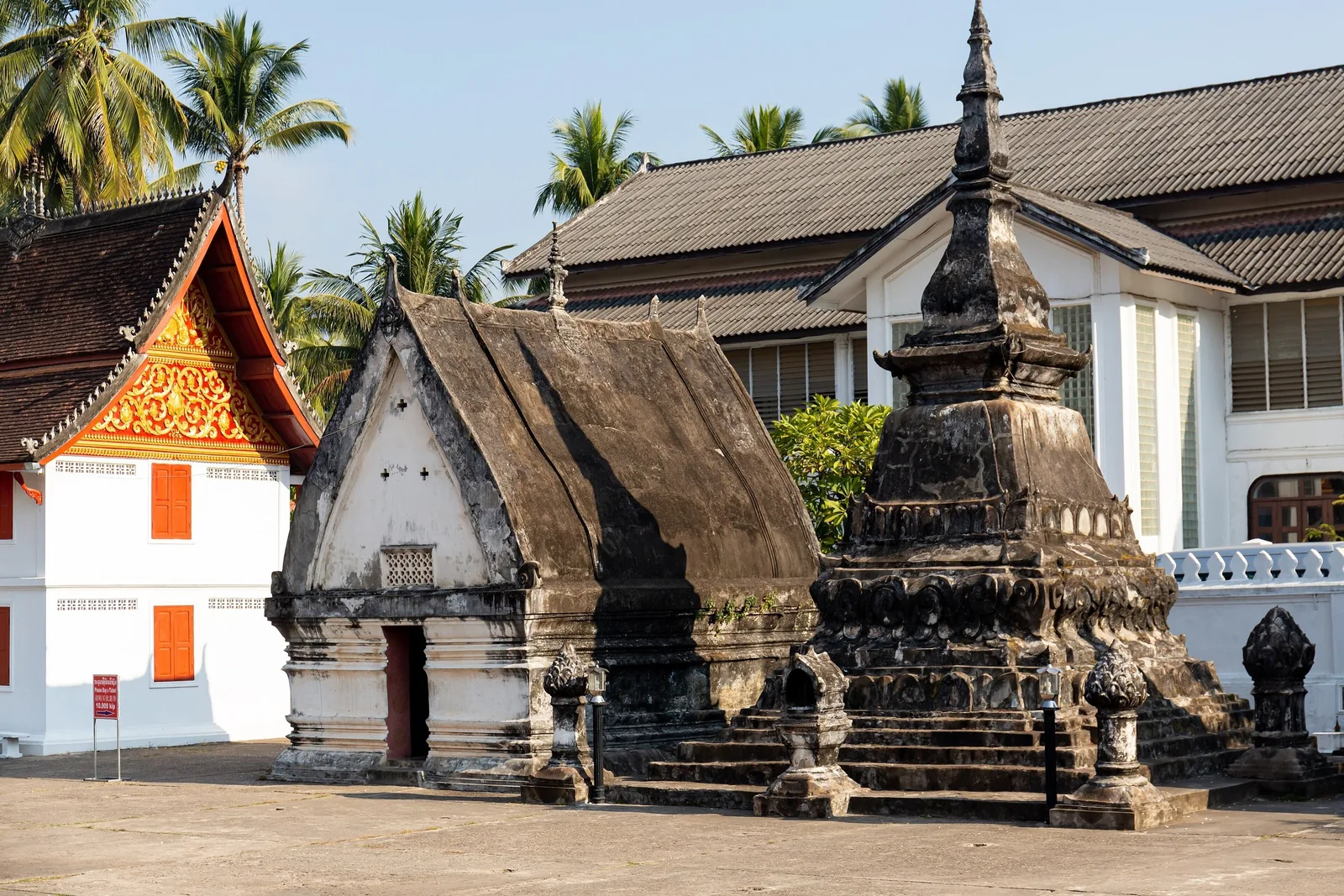 A weathered stupa and small chapel stand before white temple buildings in Luang Prabang.