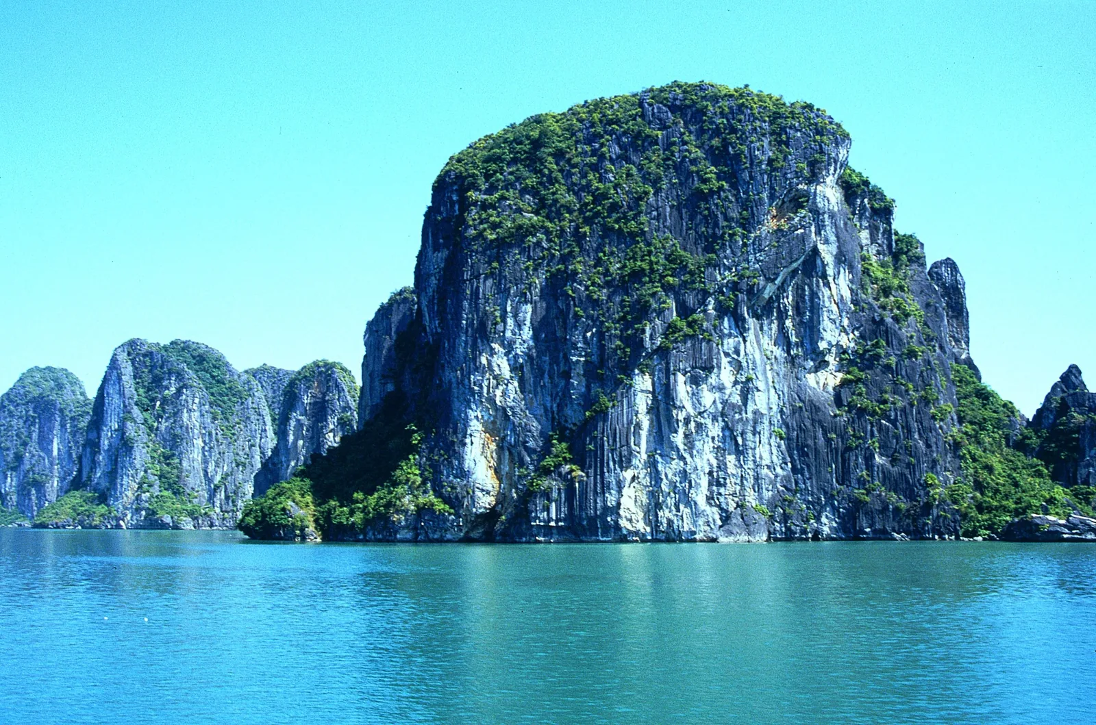 A tall limestone karst covered in greenery rises from the emerald waters of Ha Long Bay under a clear sky.