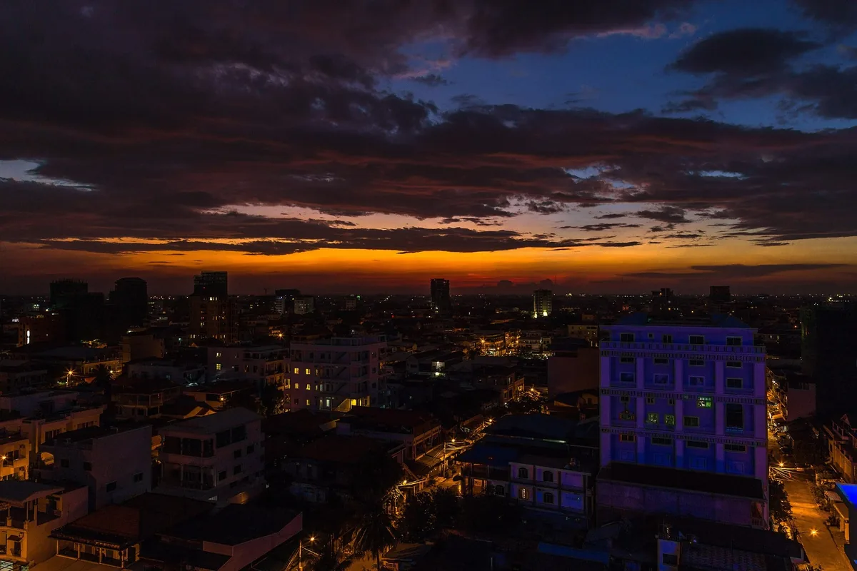 A dramatic sunset glows orange behind the darkening skyline of Phnom Penh, with city lights beginning to twinkle below.