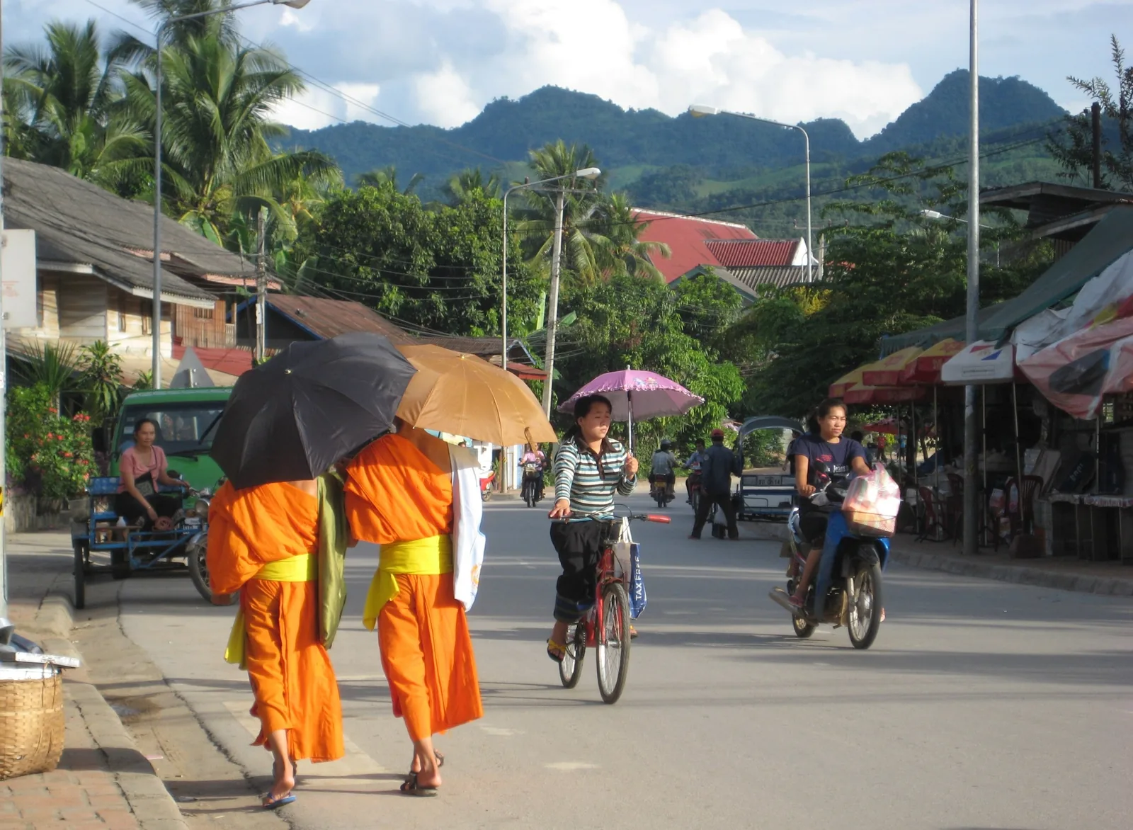 Monks in orange robes with umbrellas walk a Luang Prabang street as cyclists and mountains complete the scene.
