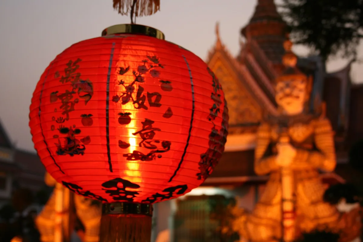 A red Chinese lantern glows in front of an illuminated temple guardian statue in Bangkok at dusk.