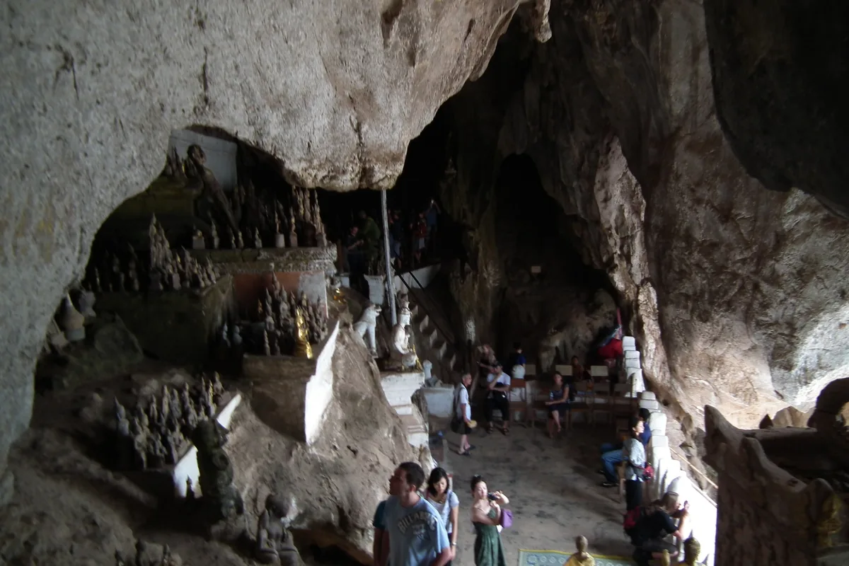 Rows of small Buddha statues fill the rocky interior of the Pak Ou Caves near Luang Prabang, with visitors exploring below.