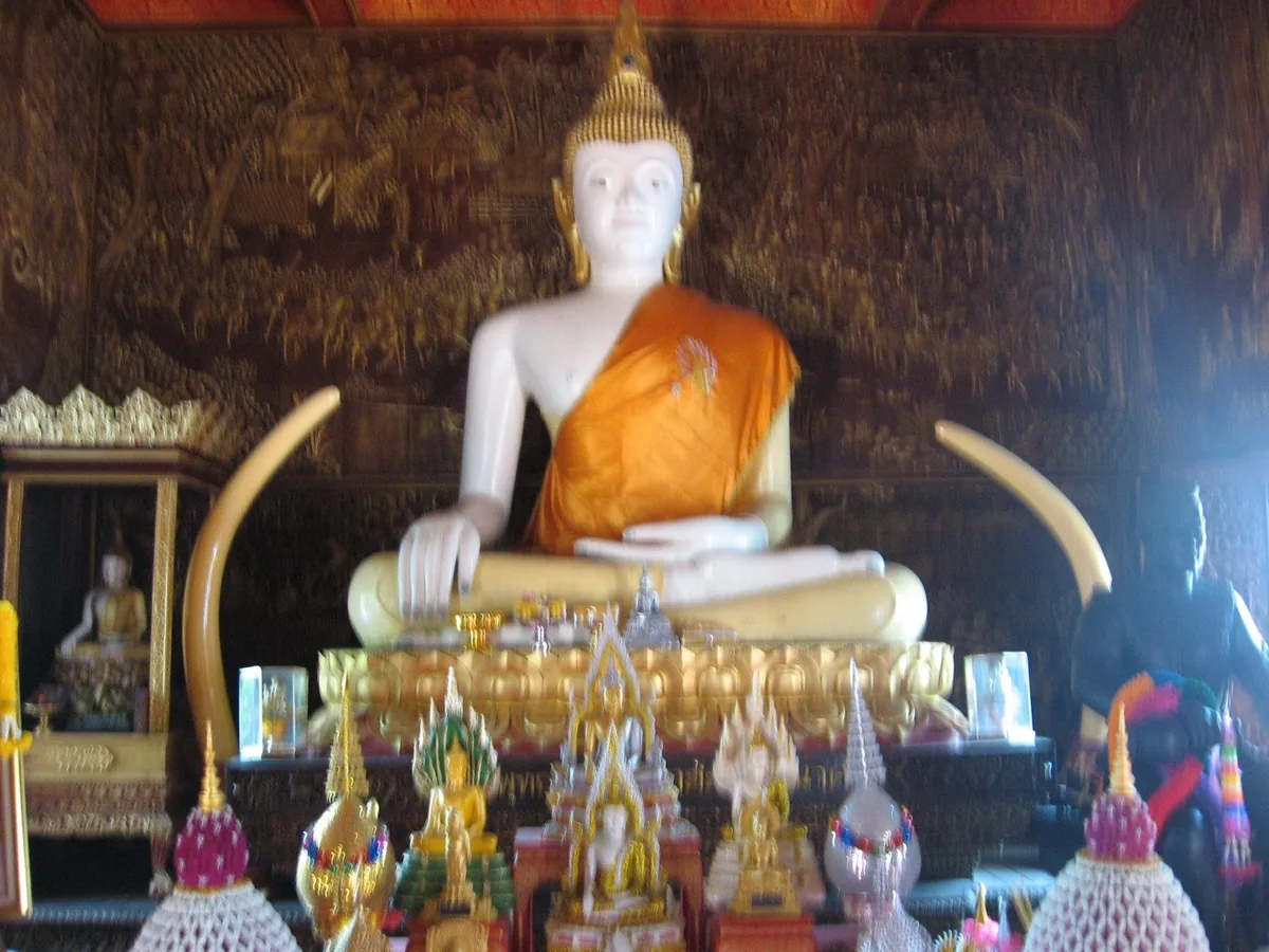 A white marble Buddha statue draped in an orange robe sits inside a temple hall in Chiang Mai, flanked by large tusks and.