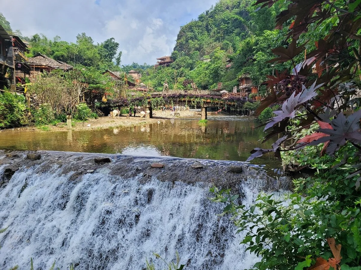 A small waterfall flows past a wooden bridge and stilt houses in Cat Cat Village, surrounded by lush green hills.