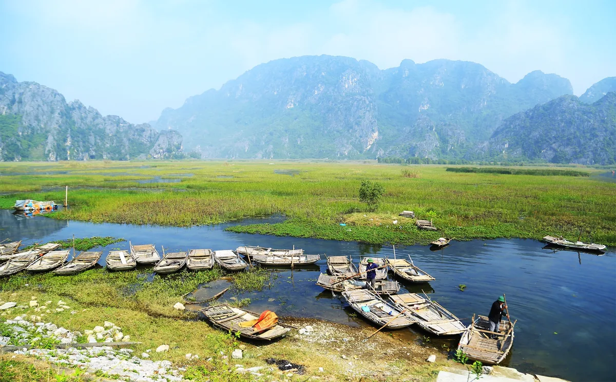 Wooden sampans dock in a marshy river channel below limestone karst mountains at Van Long, Ninh Binh.