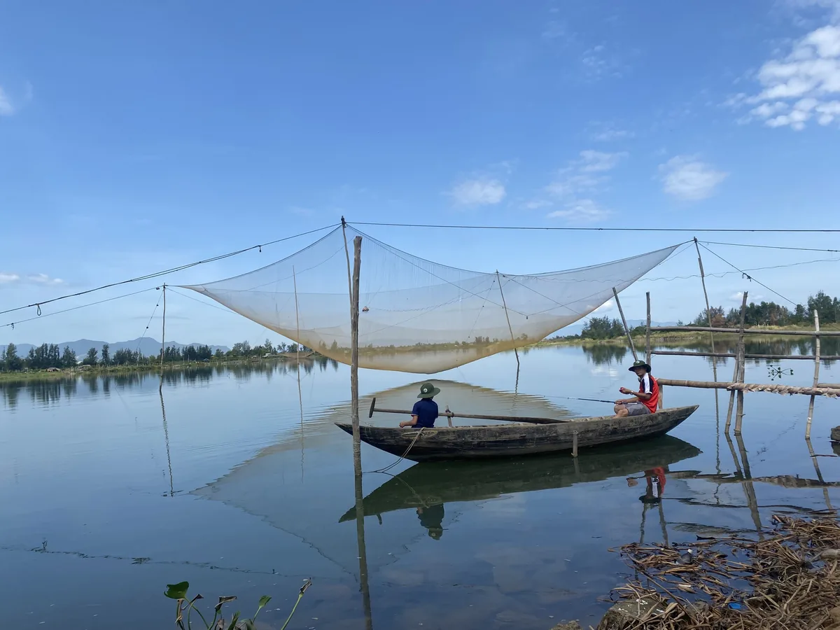 A traditional square lift net hangs over a calm river as two people fish from a wooden boat in the Hoi An countryside.