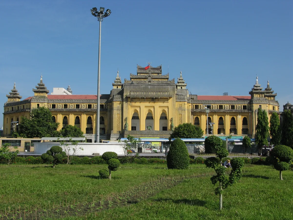Yangon City Hall's ornate yellow façade with tiered spires rises behind a manicured lawn under a clear blue sky.