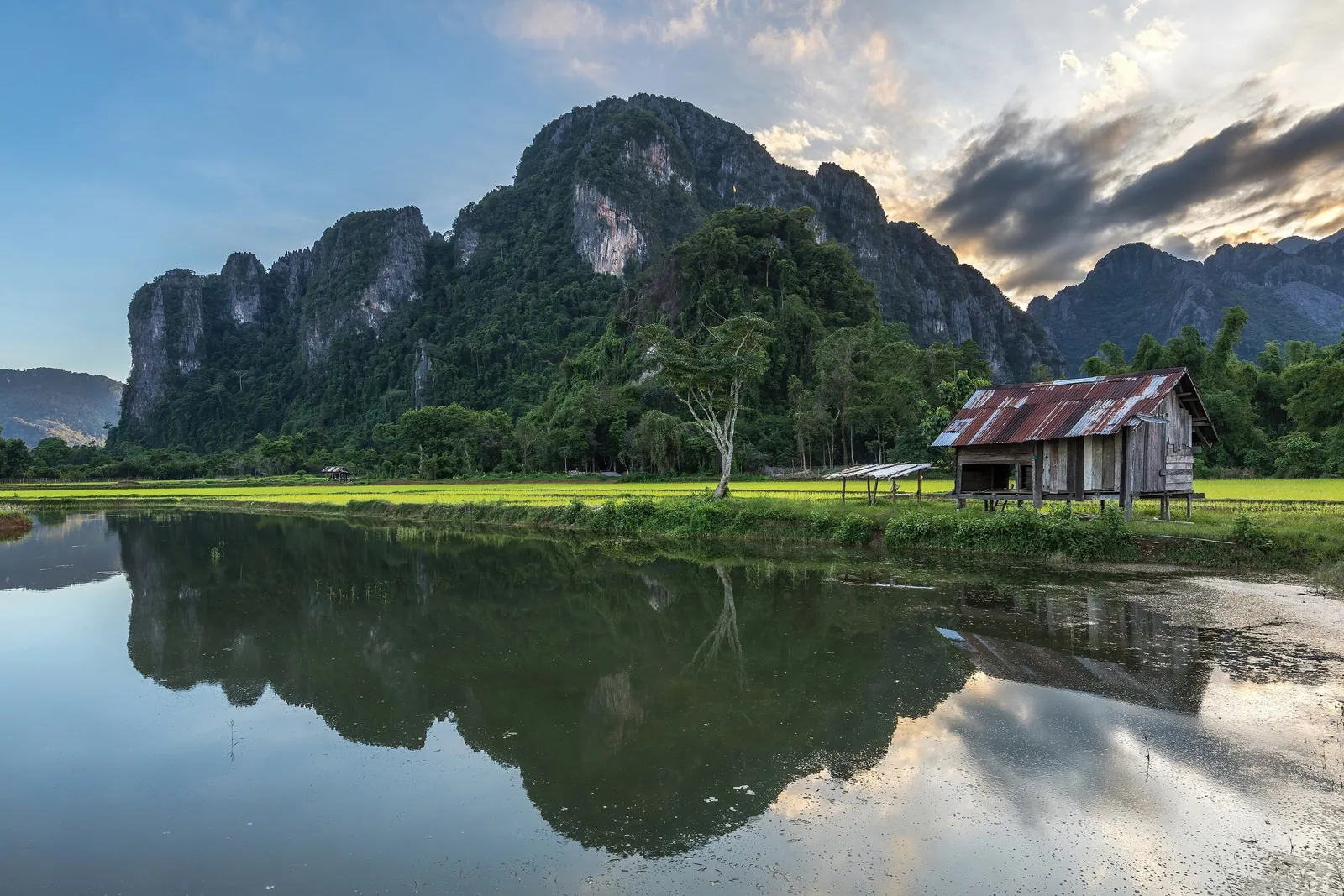 Limestone karst mountains reflect in a still rice paddy pond beside a wooden hut in Vang Vieng, Laos.