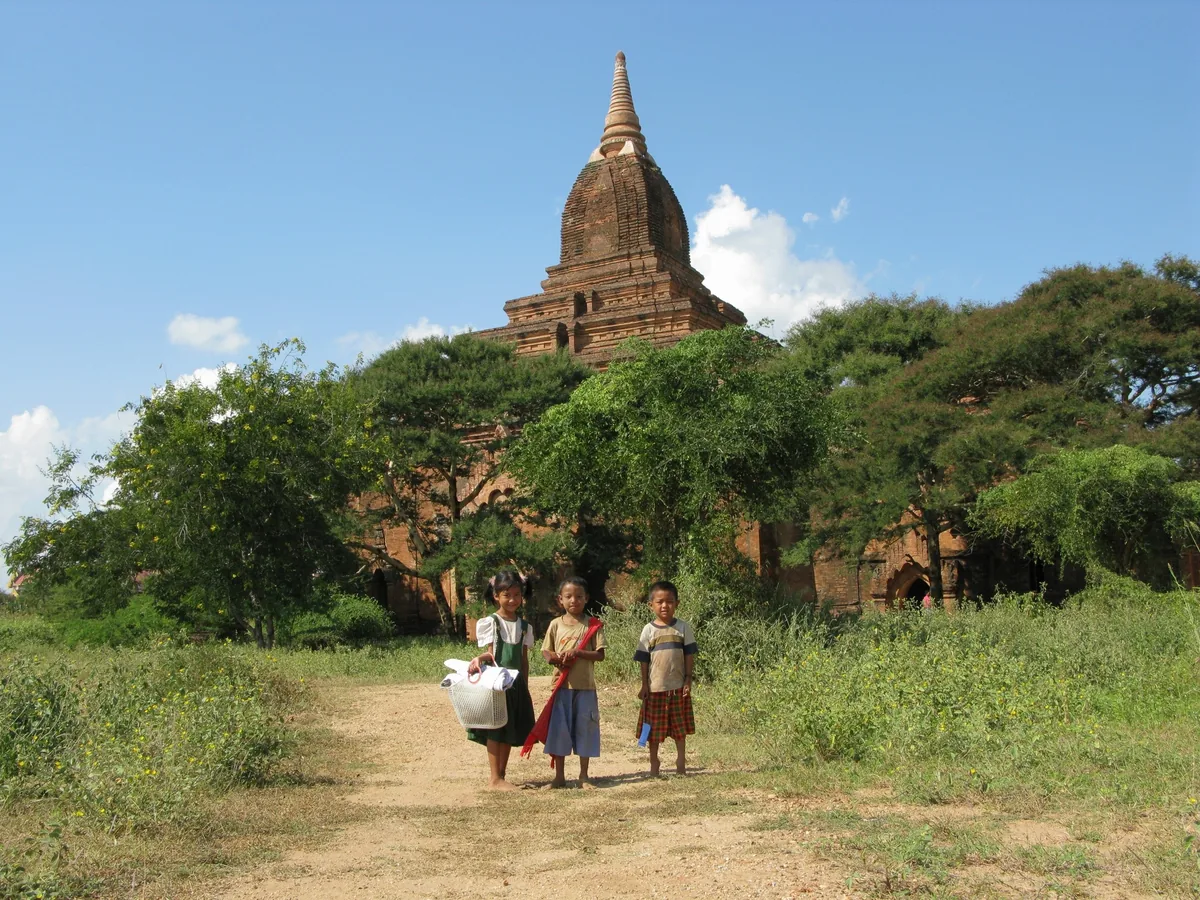 Local children stand on a dirt path near an ancient brick pagoda surrounded by trees in Bagan.