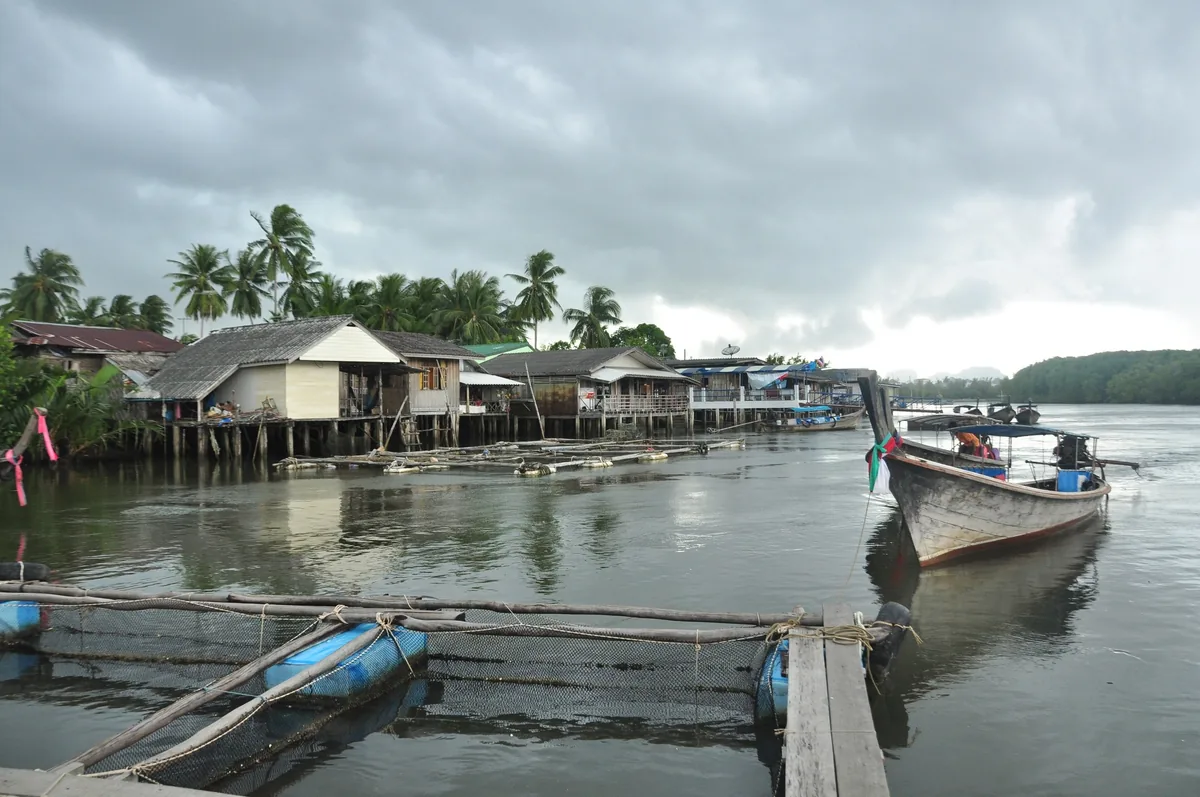 Stilt houses and longtail boats line a calm river in a fishing village near Krabi Town under cloudy skies.