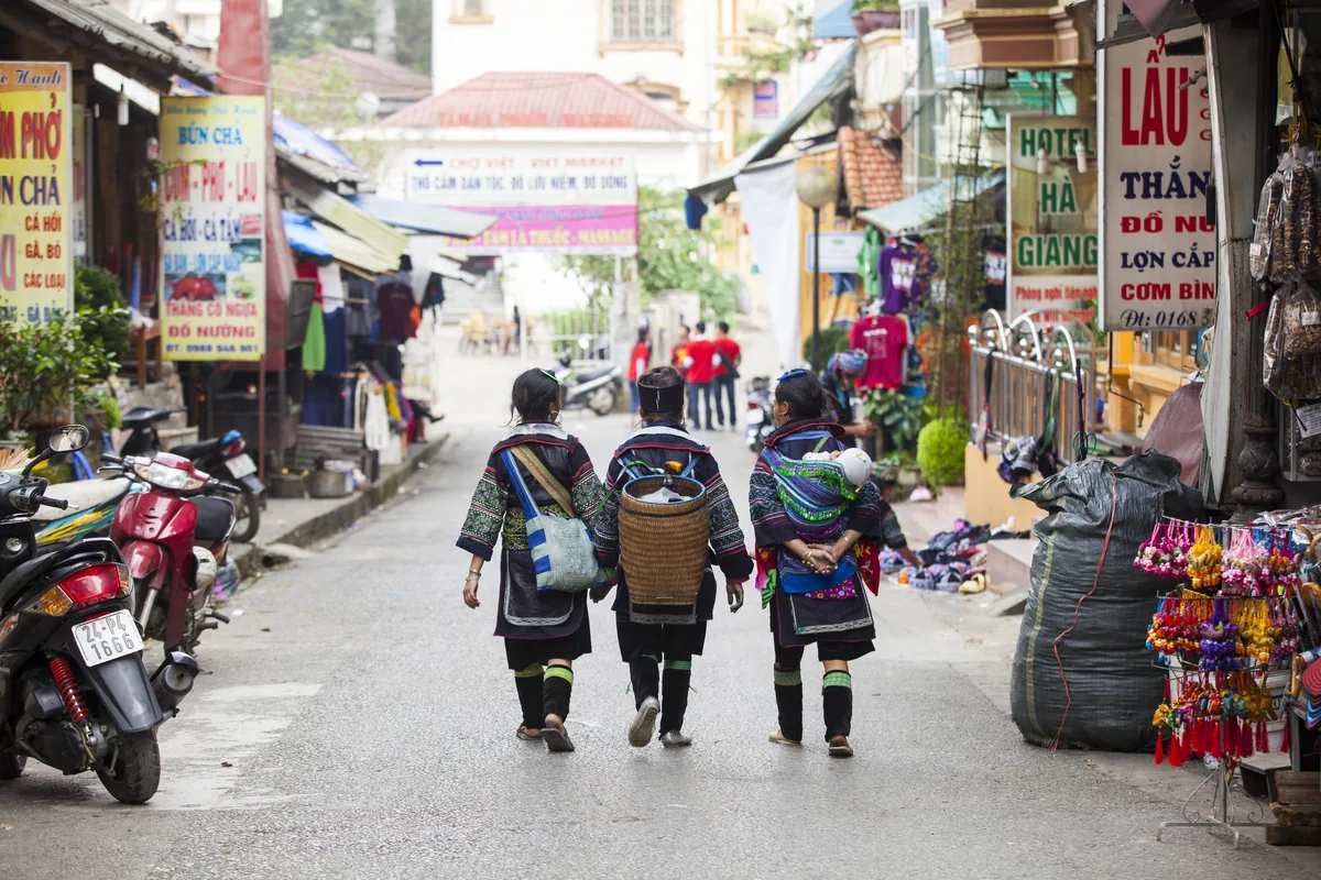 Three Hmong women in traditional dress walk down a busy street lined with shops in Sapa.