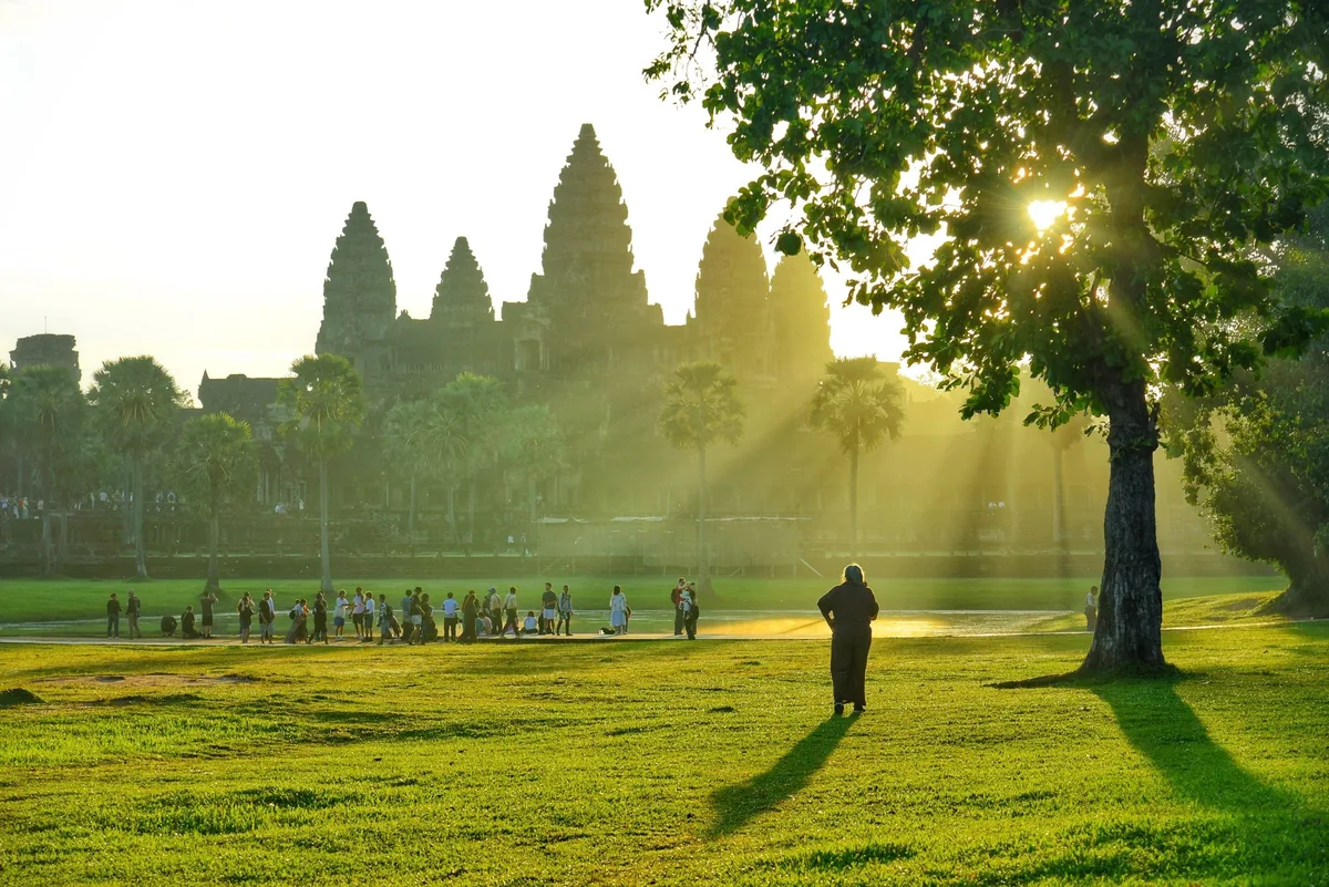 Sunlight streams through trees as visitors gather on the lawn before Angkor Wat's iconic towers at sunrise.