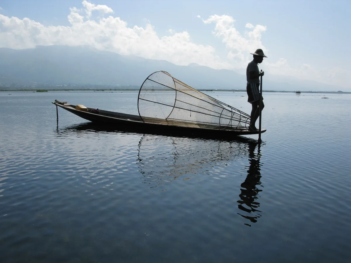 A fisherman leg-rowing his boat with a conical bamboo net on the calm waters of Inle Lake, Myanmar.