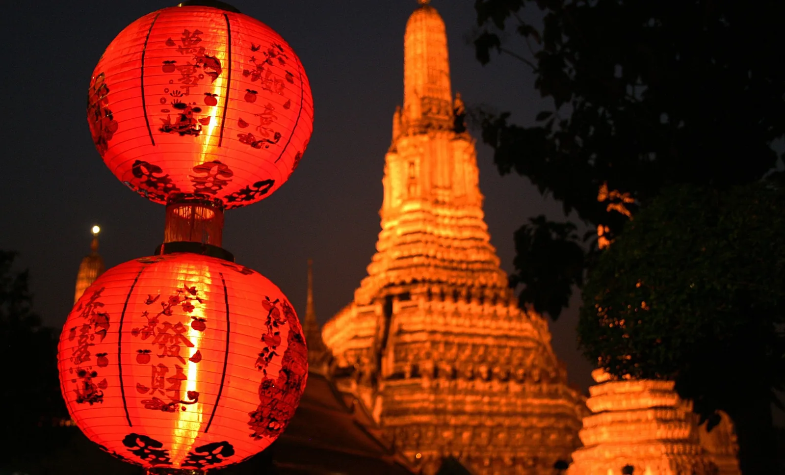 Red lanterns glow beside the illuminated spire of Wat Arun temple at night in Bangkok.