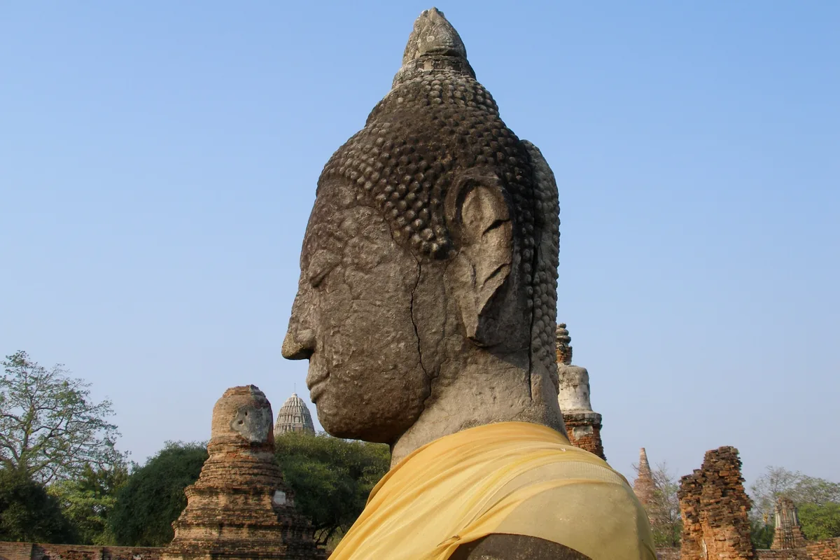 A weathered stone Buddha head draped in yellow cloth stands among ancient temple ruins in Ayutthaya, Thailand.