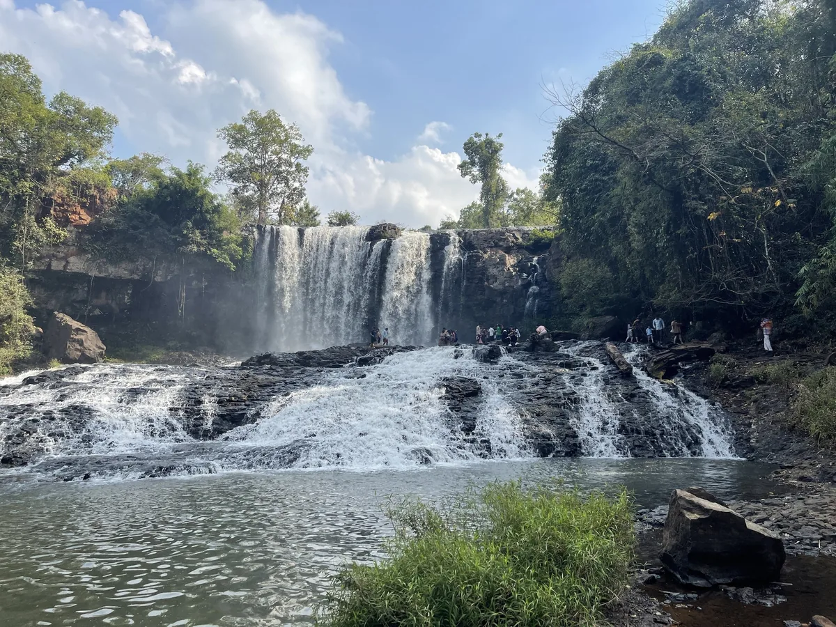 Wide cascade tumbles over dark rock ledges at Bousra Waterfall in Mondulkiri, with visitors gathered near the base.