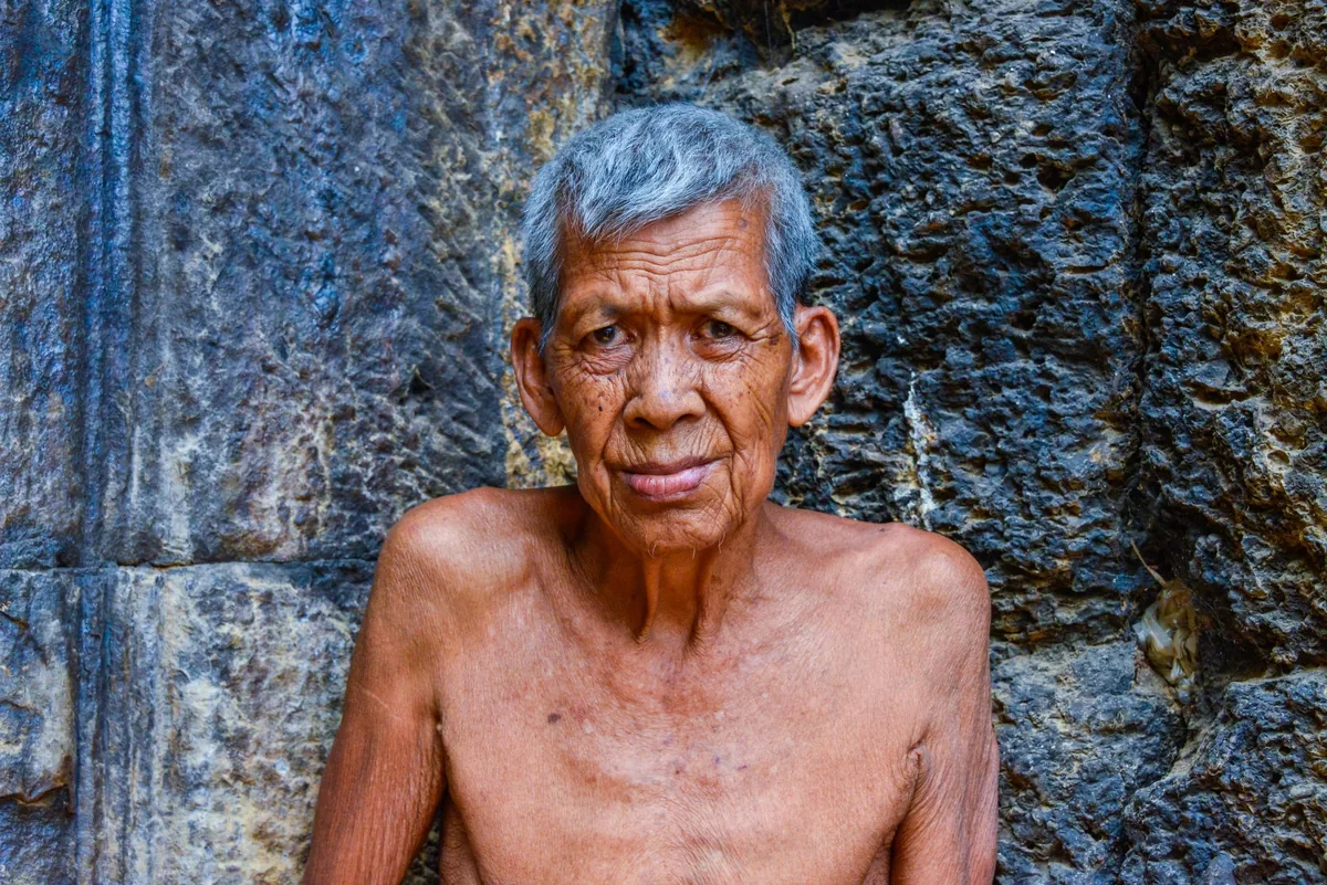 An elderly Cambodian man sits shirtless against a weathered stone wall, gazing at the camera.
