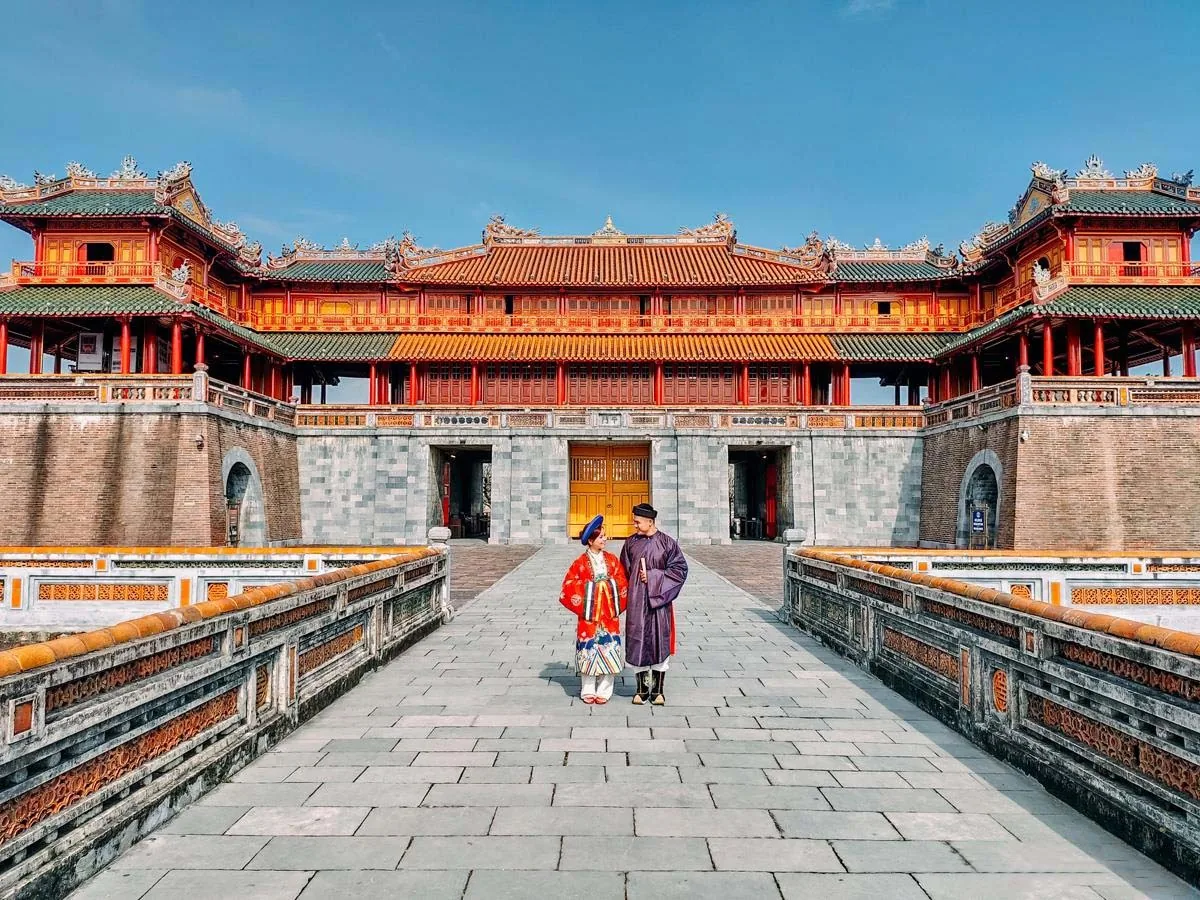 Two people in traditional imperial costumes walk across a stone bridge toward the ornate Ngo Mon Gate of the Hue Imperial.