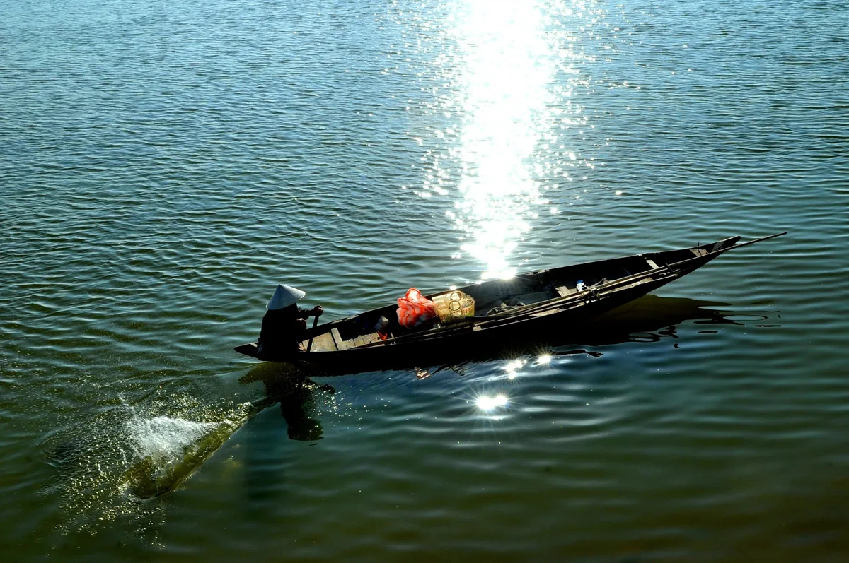 A person in a conical hat rows a small wooden boat across the sunlit Han River in Da Nang.