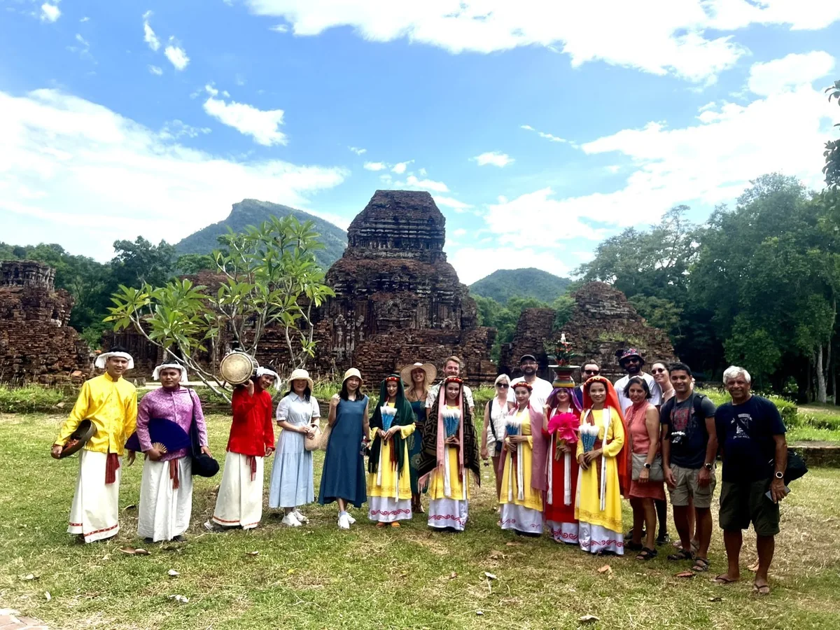Tourists and Cham dancers in traditional dress pose before the ancient brick towers of My Son Sanctuary.