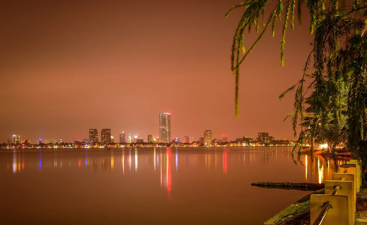 City skyline lights reflect across West Lake at night in Hanoi, framed by drooping tree branches.