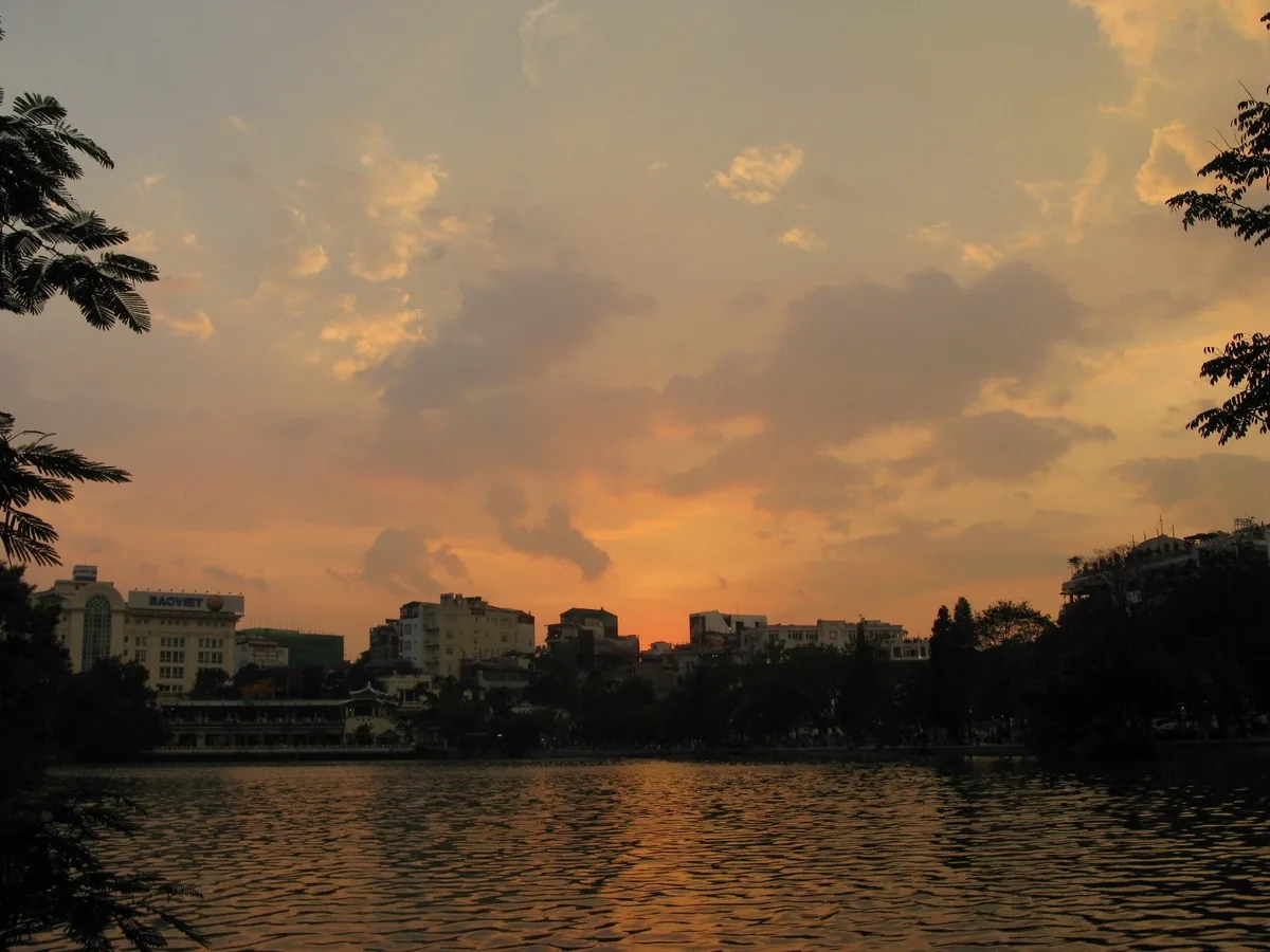 Orange sunset sky over Hoan Kiem Lake with silhouetted city buildings and trees in Hanoi
