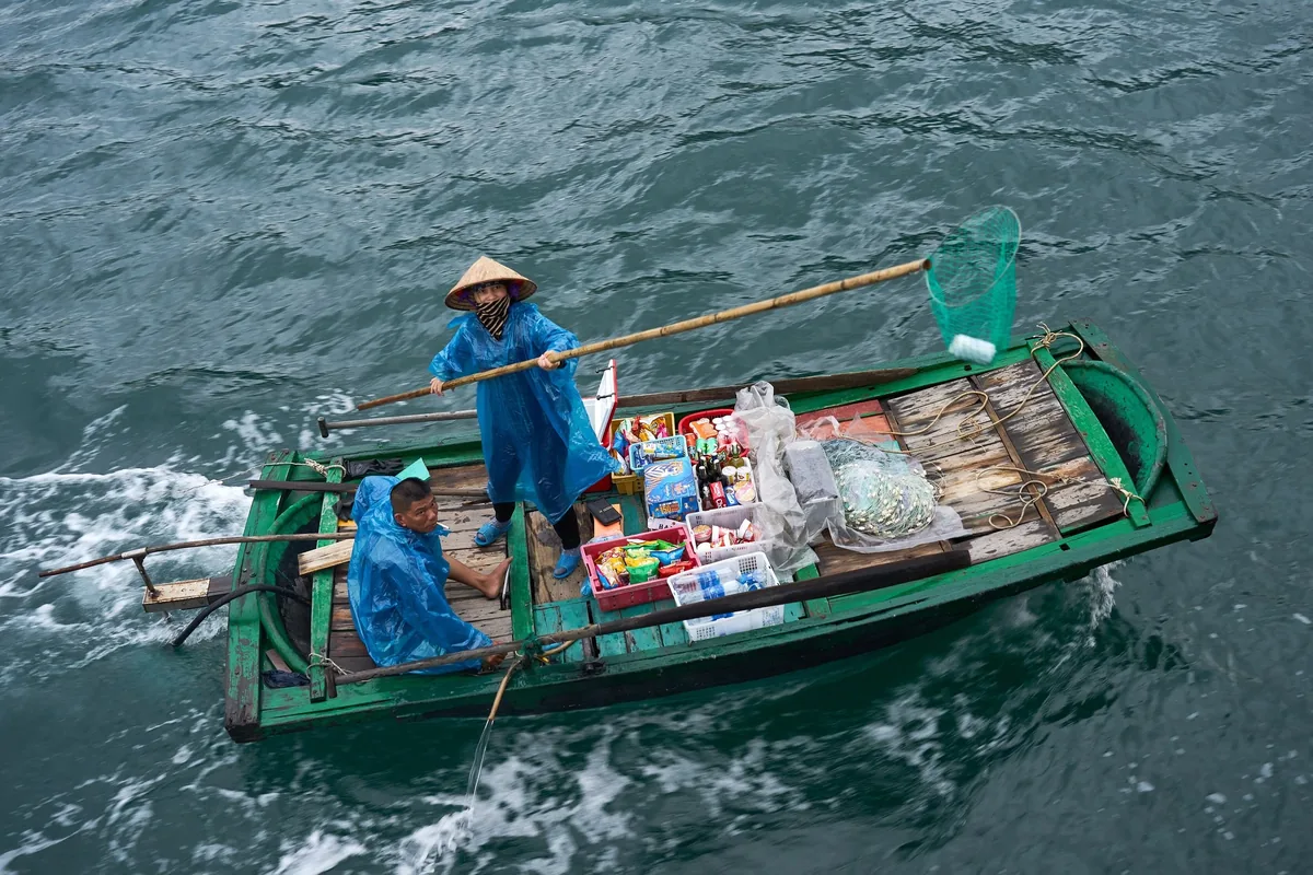 A vendor in a conical hat rows a small green boat loaded with goods across the water in Ha Long Bay.