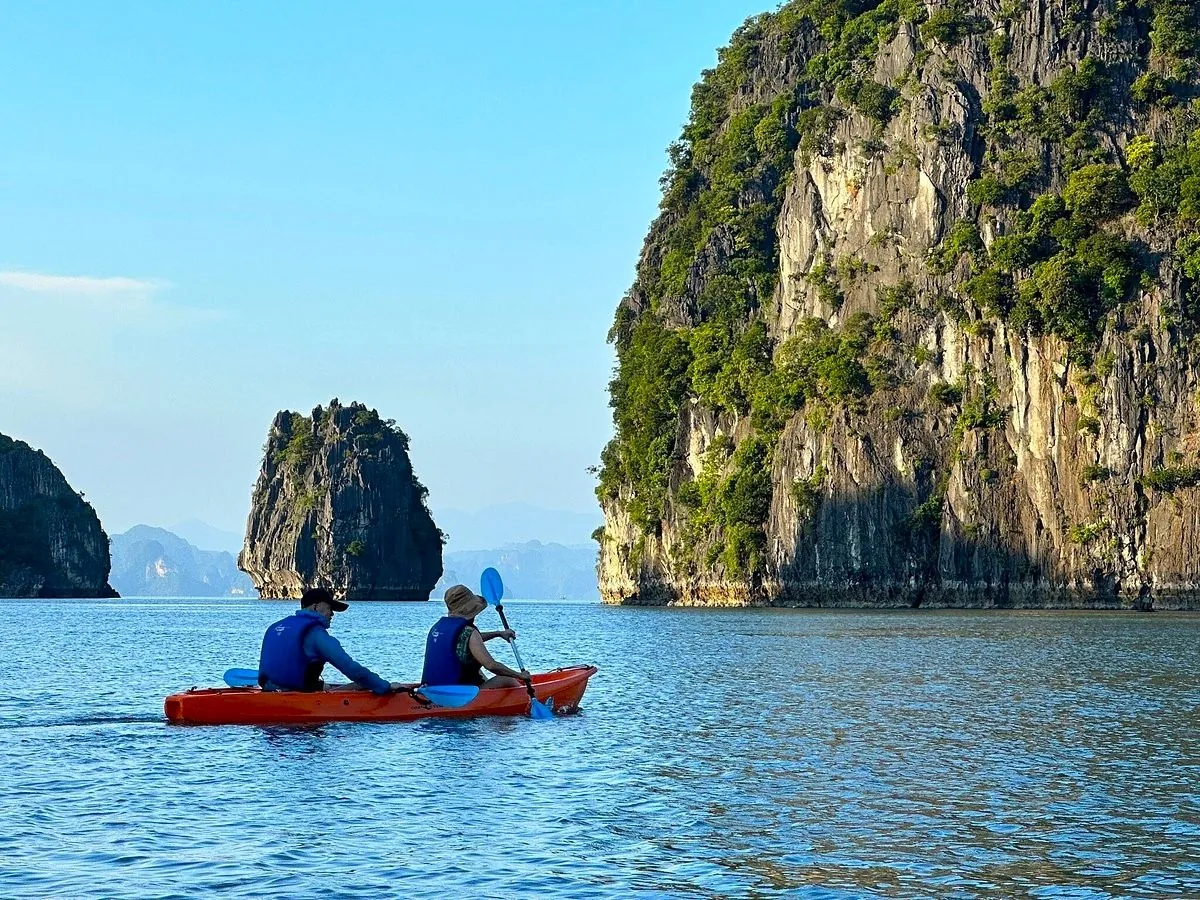 Two people paddle an orange kayak past limestone karsts in Bai Tu Long Bay.