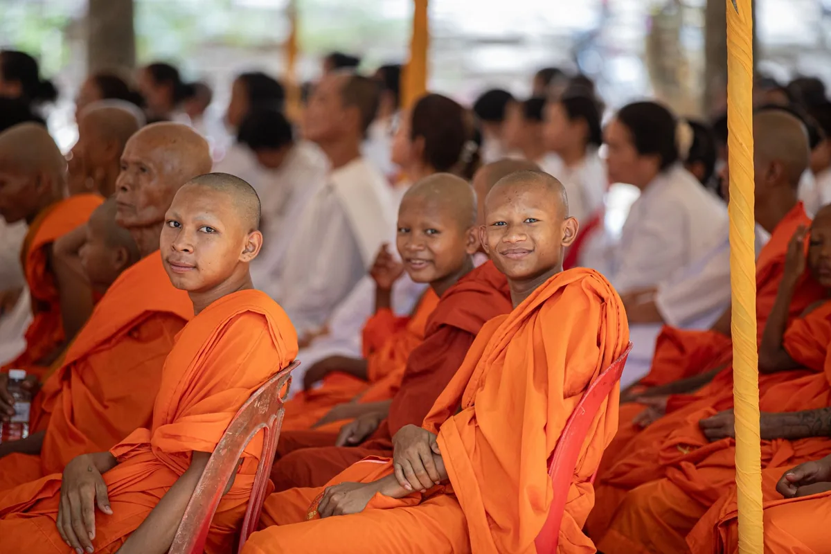 Young Buddhist novices in saffron robes sit smiling among monks and lay worshippers at a Cambodian monastery gathering.