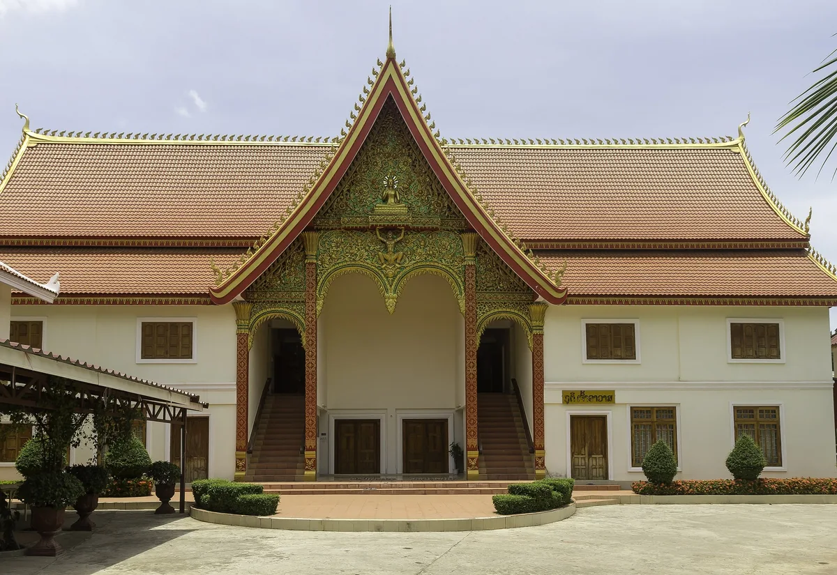 A cream-colored Buddhist temple hall with an ornate gilded gable and tiered roof stands under a bright sky in Vientiane.