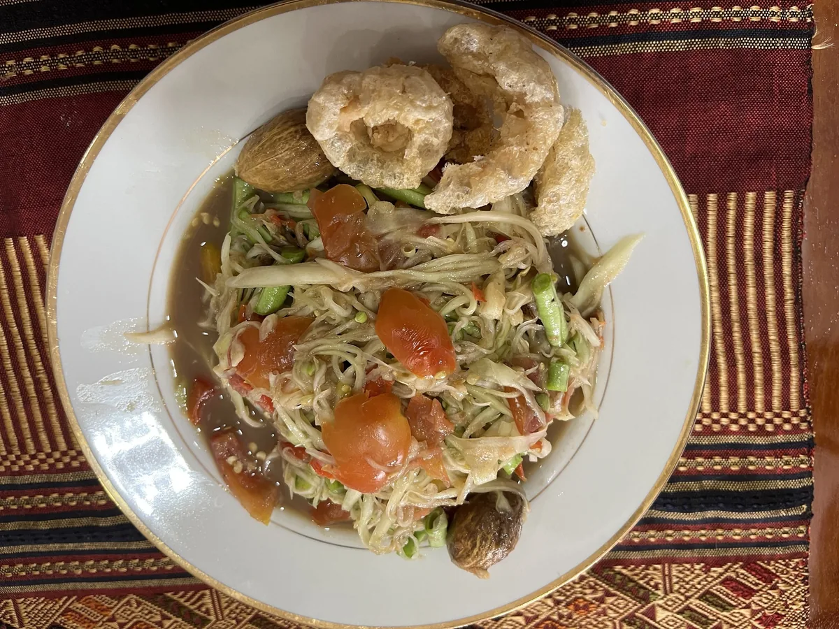 A plate of Lao papaya salad with tomatoes, herbs and crispy pork rind on a woven mat.